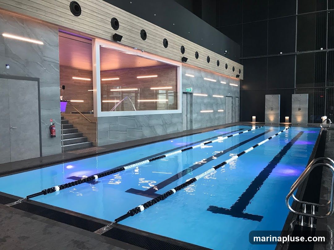 An indoor swimming pool with multiple lanes marked by black and white ropes. The pool is illuminated by blue and purple lights. The surrounding area features modern architecture with grey tiled walls, wooden accents, and sleek lighting fixtures. There are stairs leading to a higher level with a glass partition, and shower stalls are visible on the far side. A fire extinguisher is mounted on the wall near a door.
