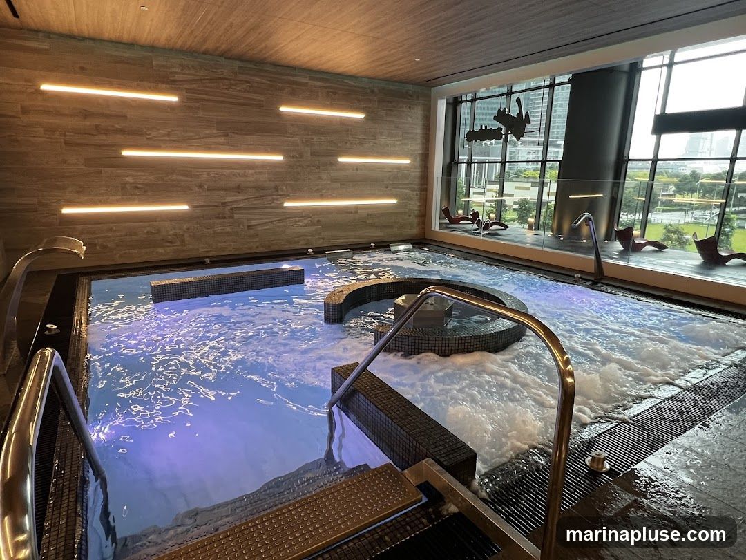 A spacious indoor jacuzzi with bubbling water and purple lighting. The jacuzzi has tiled steps and metal railings. Large windows overlook a cityscape with modern buildings and green spaces. Several lounge chairs are visible outside the windows. The walls are decorated with horizontal wooden panels and linear lights.