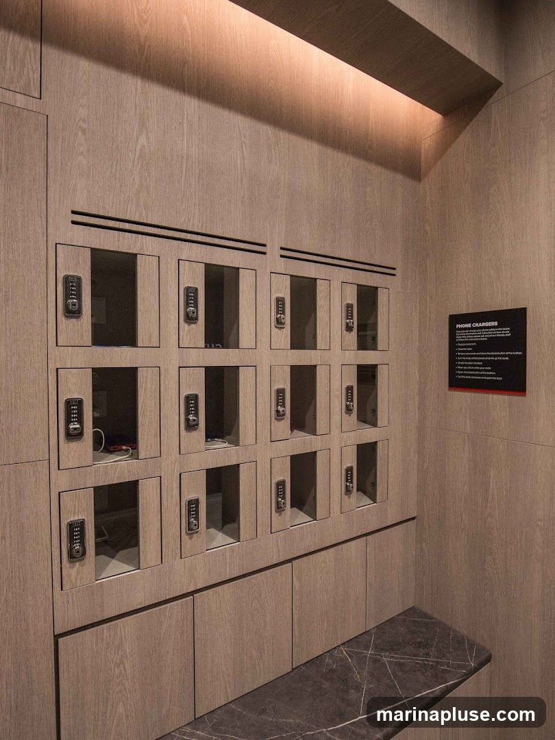 A wall of wooden lockers with digital keypads, designed for charging phones. Some lockers are open, revealing charging cables and devices inside. A dark marble bench is positioned in front of the lockers. A black sign with red text titled "PHONE CHARGERS" is mounted on the wall to the right, providing instructions.