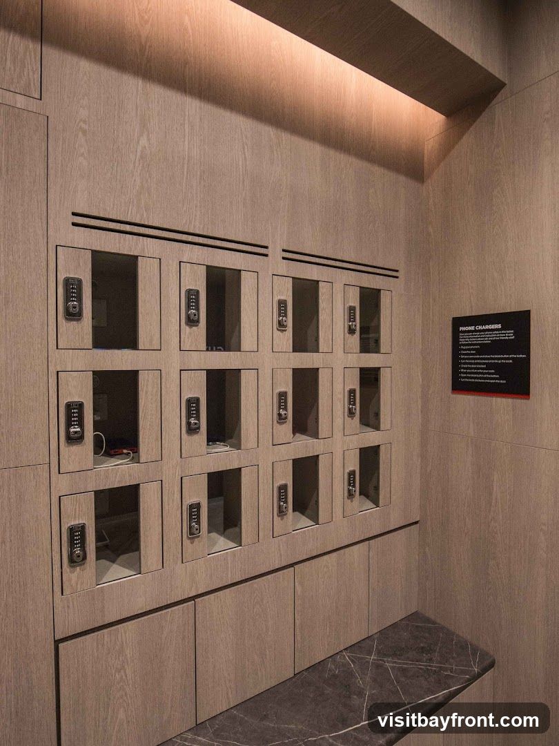 A wall of wooden lockers with digital keypads, designed for charging phones. Some lockers are open, revealing charging cables and devices inside. A dark marble bench is positioned in front of the lockers. A black sign with red text titled "PHONE CHARGERS" is mounted on the wall to the right, providing instructions.