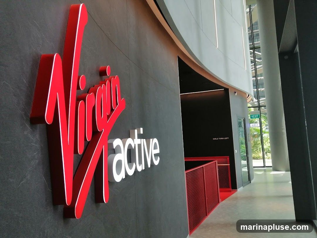 A close-up view of the Virgin Active logo, a large red script font for 'Virgin' and white block letters for 'active', mounted on a dark textured wall. To the right, a hallway leads towards an exit sign and a door labeled 'GIRLS'. Red railings are visible along a curved wall.