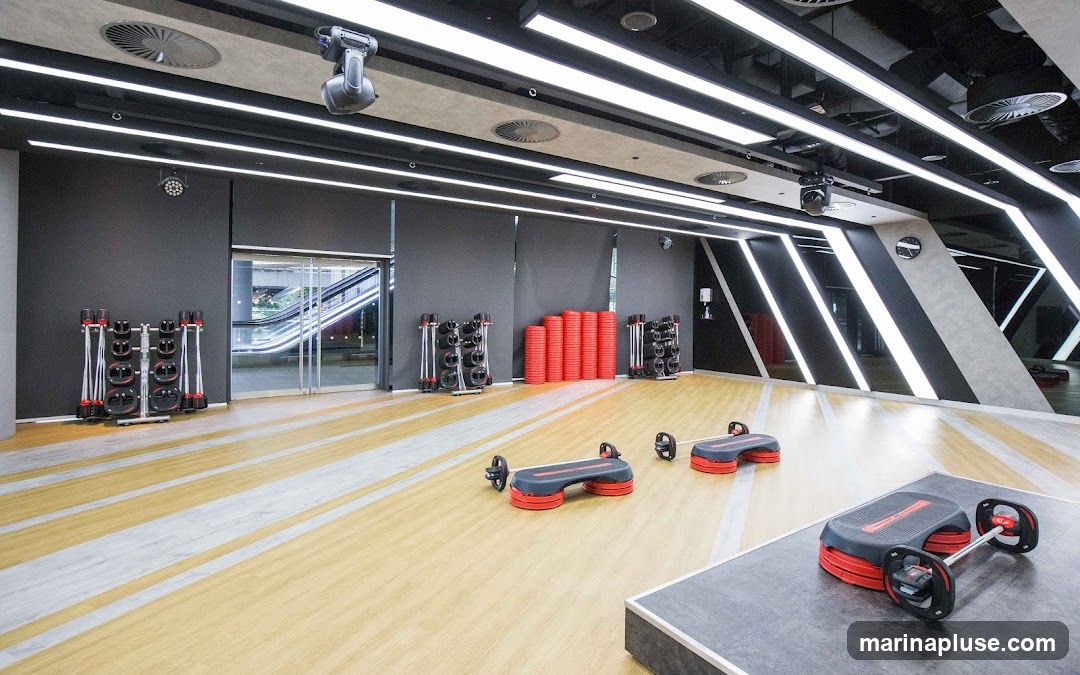 A modern gym interior with a wooden floor marked with light-colored stripes. On the left, racks of dumbbells are visible. In the center, stacks of red aerobic steps are arranged. In the foreground, two aerobic step platforms with attached barbells are positioned. The ceiling features contemporary lighting and ventilation systems. Through glass doors on the left, an escalator is visible.