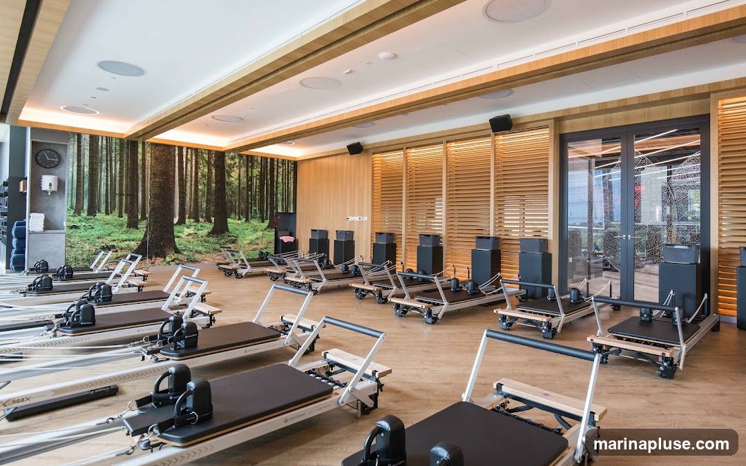 A well-lit Pilates studio with multiple reformers arranged in rows. The room features a large mural of a forest on one wall, and wooden slatted panels on others. There are speakers mounted on the ceiling and a clock on the wall. The floor is light wood.