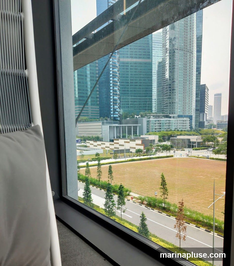 A view of a modern cityscape with numerous tall glass buildings, seen through a large window. In the foreground, a portion of a white chair with a grey cushion is visible. Outside the window, there is a large, dry grassy field bordered by a road and lined with trees. The sky is overcast.