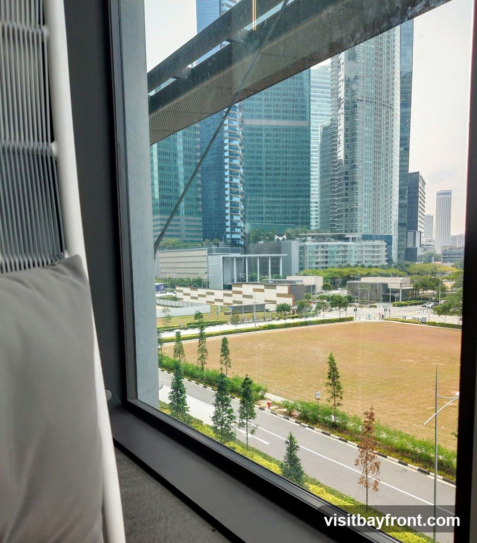 A view of a modern cityscape with numerous tall glass buildings, seen through a large window. In the foreground, a portion of a white chair with a grey cushion is visible. Outside the window, there is a large, dry grassy field bordered by a road and lined with trees. The sky is overcast.