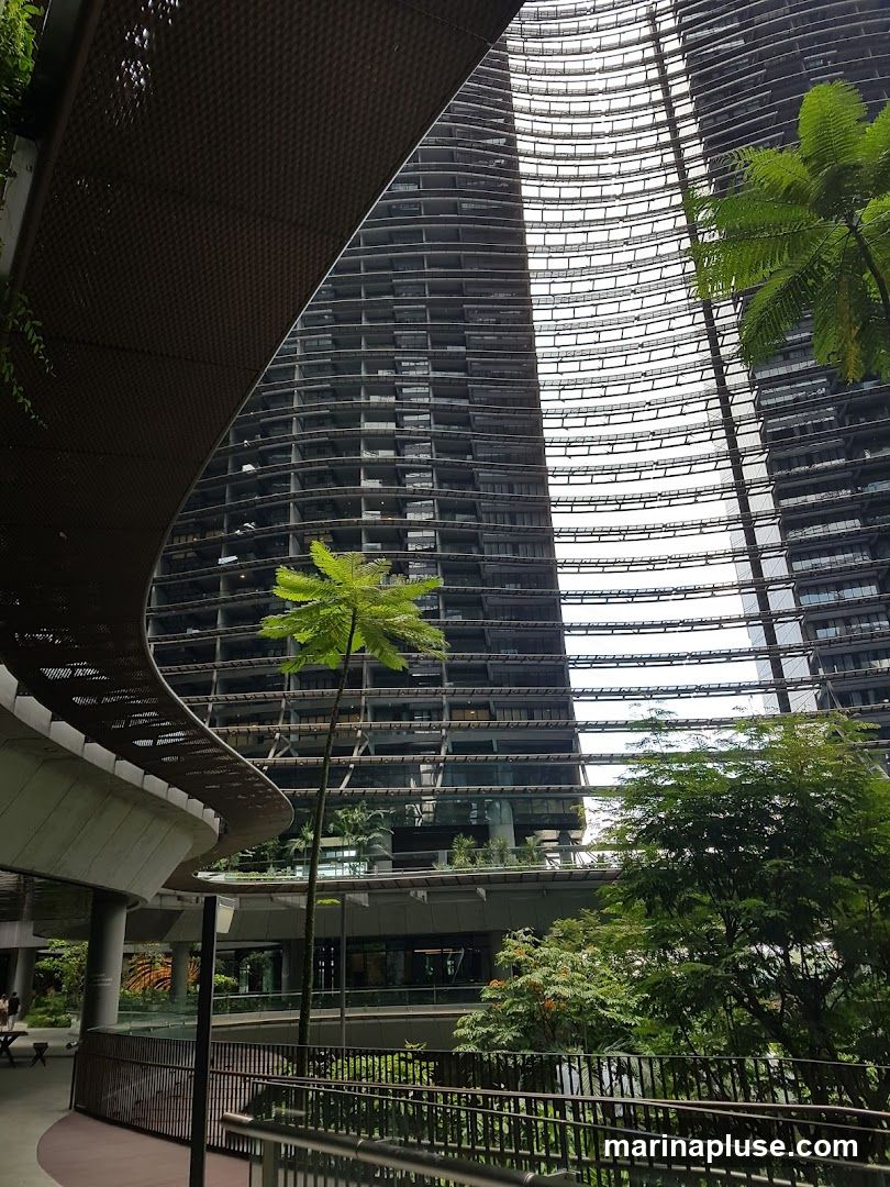 A view looking up at the striking, curved facades of modern skyscrapers, interspersed with lush green trees and foliage. A walkway with a metal railing is visible in the foreground, leading towards the buildings. The architecture features a repeating pattern of horizontal lines and balconies, creating a sense of depth and scale. The overall scene blends urban design with natural elements.