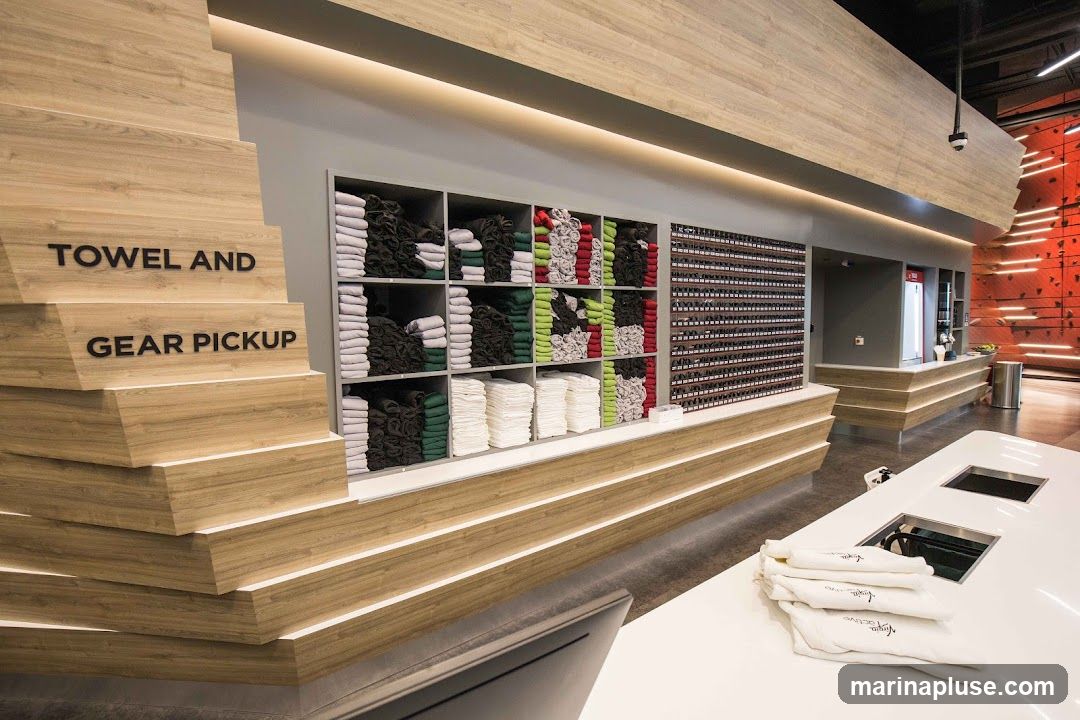 A modern interior space with a prominent wooden structure featuring the words "TOWEL AND GEAR PICKUP" in black lettering. Shelves are stocked with neatly folded towels in various colors, including white, grey, dark green, and red. To the right, a wall is covered with numerous small items, possibly lockers or storage compartments. In the background, a counter area is visible with a refrigerator and a climbing wall with illuminated orange accents. A white counter in the foreground displays folded white towels with a logo.