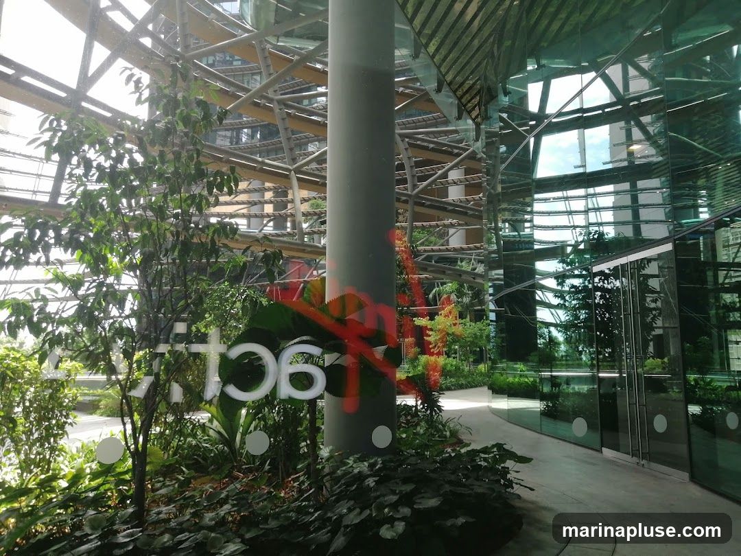 An interior view of a modern building atrium featuring a striking architectural design with exposed metal beams and a glass facade. Lush green plants and trees are strategically placed throughout the space, creating a natural and serene ambiance. The reflective glass surfaces mirror the surrounding structures and the sky, adding depth and visual interest. A large cylindrical pillar stands prominently in the foreground, partially obscured by foliage. Red graffiti is visible on the pillar and some of the glass.