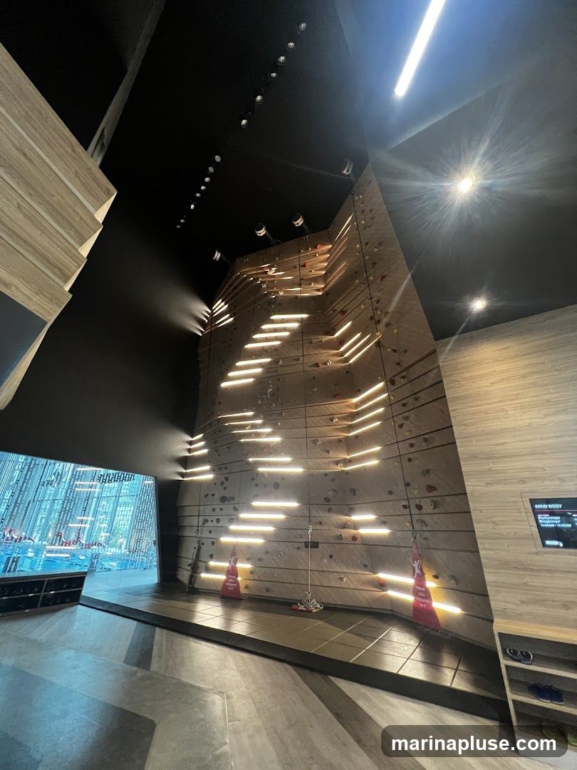 An indoor climbing wall with numerous holds and ledges. The wall is illuminated by horizontal LED lights that create a striking visual effect. In the foreground, there is a dark floor with a geometric pattern. To the left, a large window offers a view of an outdoor area with a swimming pool. To the right, a wooden panel features a digital screen displaying text and graphics. Shoe racks are visible on the left and right sides of the image.