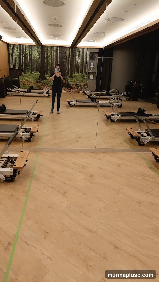 A person is taking a mirror selfie in a Pilates studio. The studio has a wooden floor with green tape markings. Several Pilates reformer machines are neatly arranged in rows. The back wall features a scenic mural of a forest. The room is well-lit with modern ceiling lights and has large mirrors on one side reflecting the machines and the person. A clock is visible on the wall.