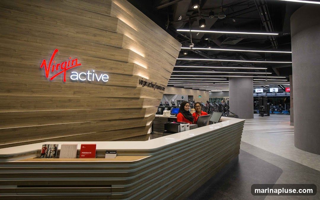 A modern gym reception area with a prominent "Virgin Active" logo on a wooden paneled wall. Two receptionists are visible behind a curved counter, attending to computers. The gym floor with exercise equipment is visible in the background.