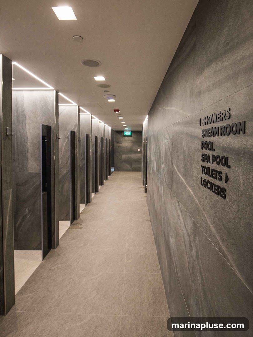 A long hallway with rows of doors on the left and a stone-tiled wall on the right. The wall has black lettering indicating directions to showers, steam room, pool, spa pool, toilets, and lockers. An illuminated green exit sign is visible in the distance.