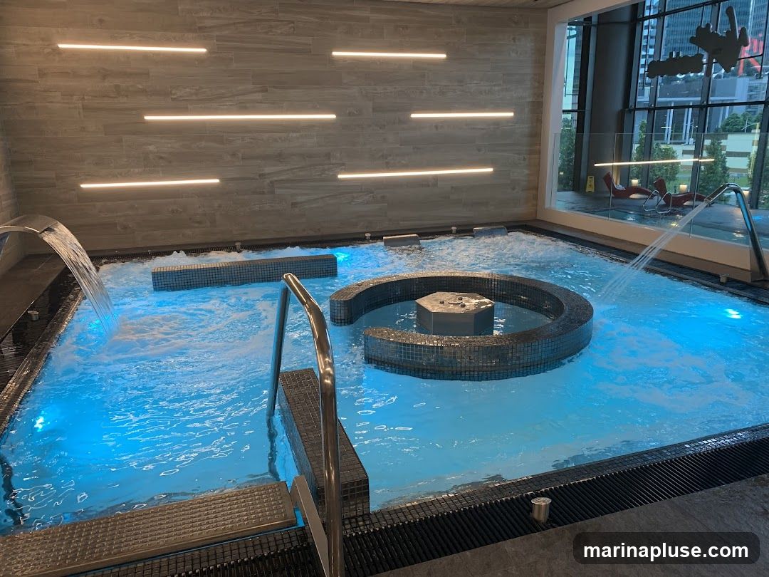 An indoor hydrotherapy pool with blue illuminated water, featuring jets, a central circular seating area with a hexagonal water feature, and a tiled wall with horizontal LED lighting. A large window overlooks an urban cityscape and lounge chairs.