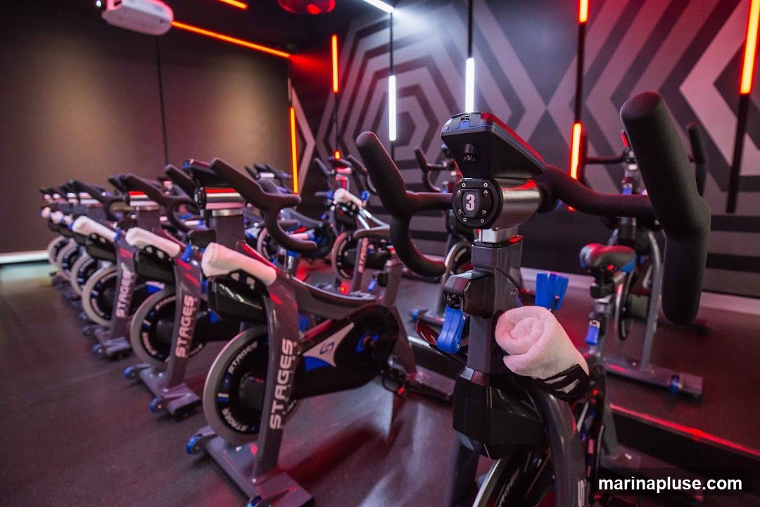 A row of stationary bicycles in a dimly lit indoor cycling studio. The bikes are dark gray with blue accents and have the brand name "Stages" visible on the frames. Each bike has a digital display and adjustable handlebars. The room is decorated with geometric patterns on the wall and illuminated by red and white LED lights. A projector is mounted on the ceiling. A rolled-up towel is placed on the handlebars of the closest bike.