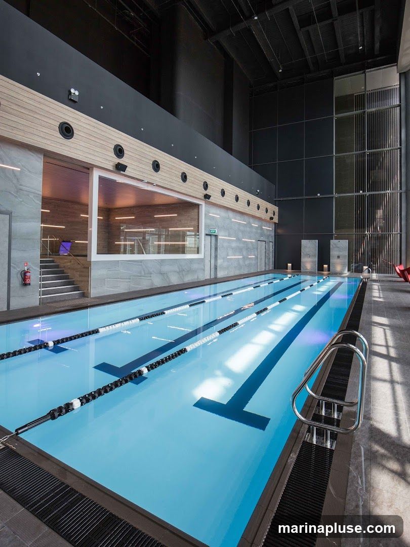 A modern indoor swimming pool with multiple lanes marked by black and white buoys. The pool has bright blue water and is surrounded by a dark tiled deck. Above the pool, there is a section with wooden paneling and a large window overlooking an interior space. The ceiling is high with exposed industrial elements. Natural light streams in from large windows on the right side of the image.