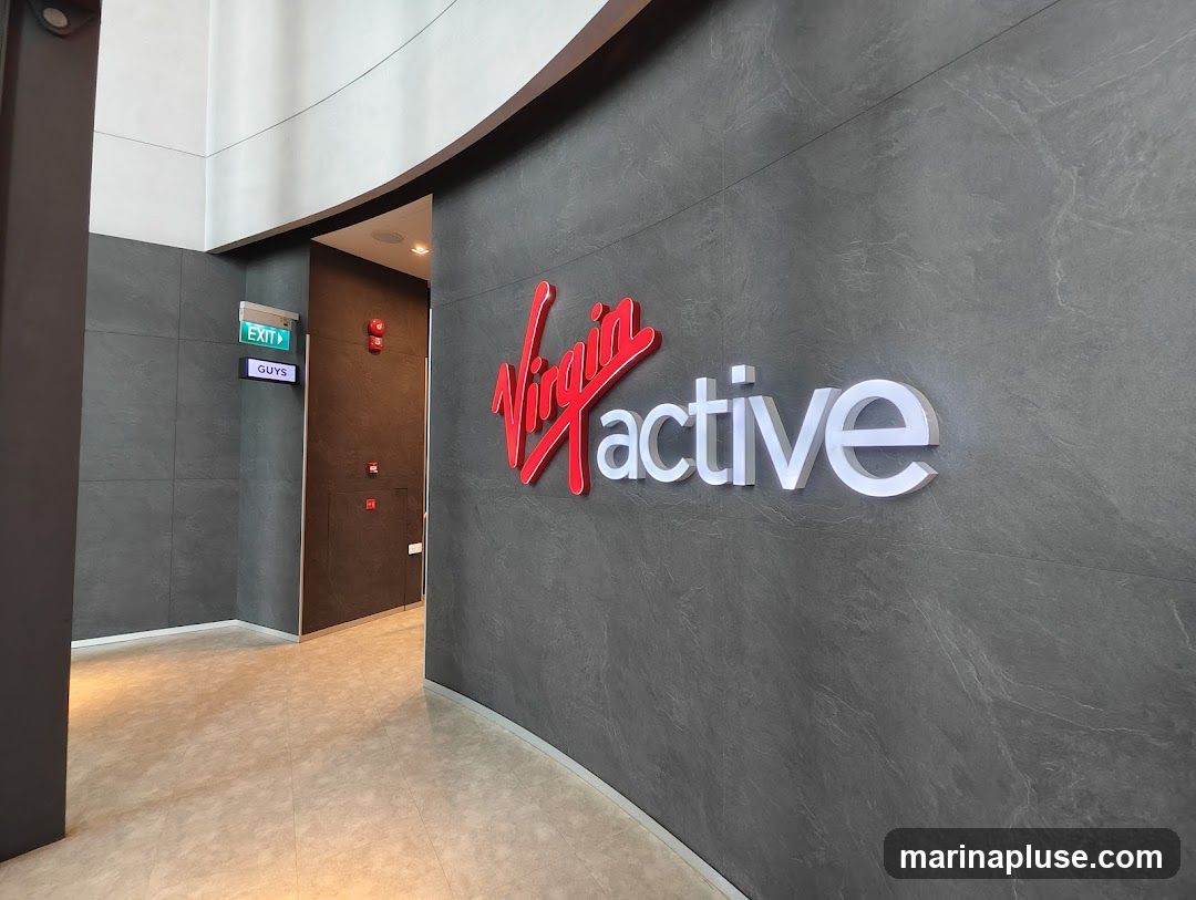 A large, illuminated sign with the Virgin logo in red and the word "active" in white is mounted on a dark, textured wall. To the left, an illuminated green exit sign is visible above a smaller sign that reads "GUYS". The floor is tiled with a light-colored pattern.