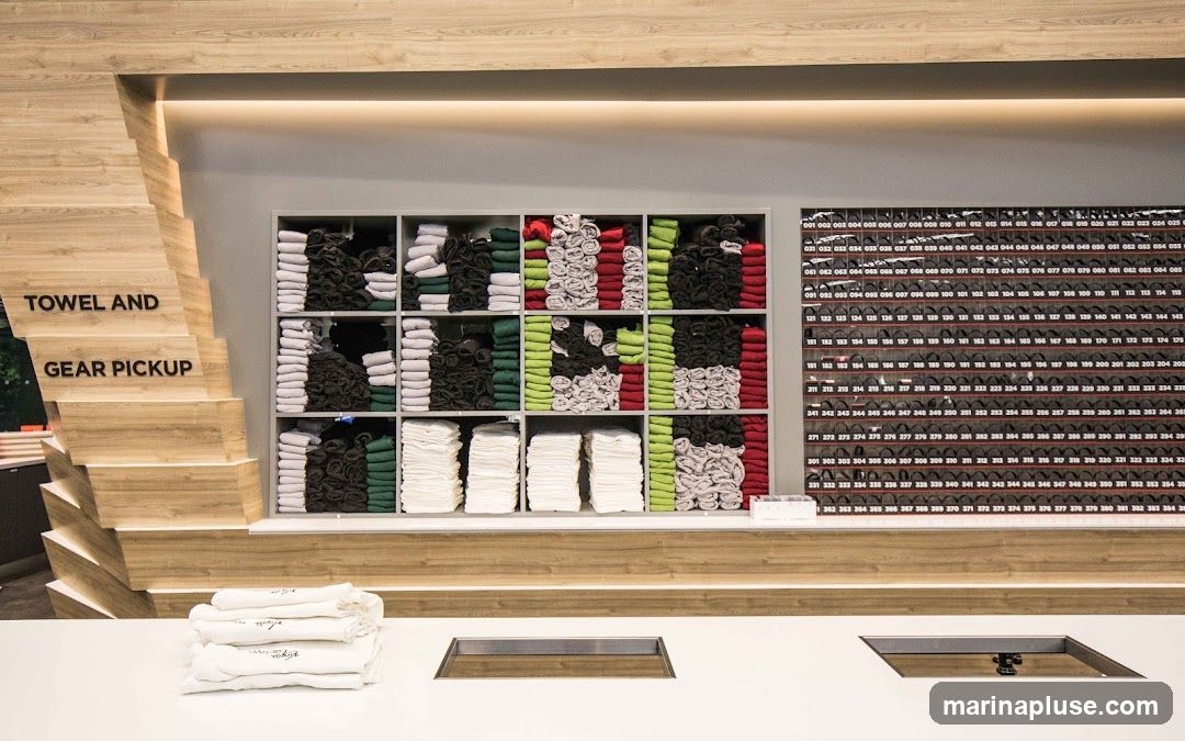 A counter with stacks of folded white towels in the foreground. Behind the counter, shelves are filled with neatly folded towels and apparel in various colors including white, black, green, and red. To the right, a wall features rows of numbered compartments, likely for storing gear. Text on a wooden structure to the left reads "TOWEL AND GEAR PICKUP.".