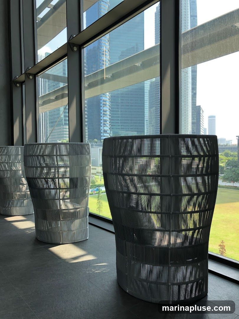 A contemporary interior space with large windows offering a panoramic view of a bustling city skyline. Three large, decorative woven planters are positioned in the foreground, casting shadows on the dark tiled floor. The planters have a unique, curved silhouette and are made of a woven material. Sunlight streams through the windows, creating a bright and airy atmosphere. The reflection in the windows shows more of the interior structure and the city outside.