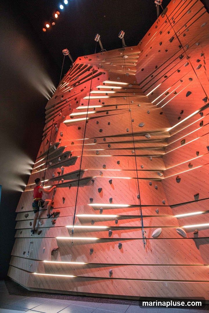 A person is climbing a large, modern indoor climbing wall. The wall is made of wood panels and features numerous climbing holds. Integrated LED lights create a dramatic effect, casting shadows and highlighting the climbing routes. The climber is wearing a red shirt and climbing gear. The background is dark, with spotlights illuminating the wall from above.