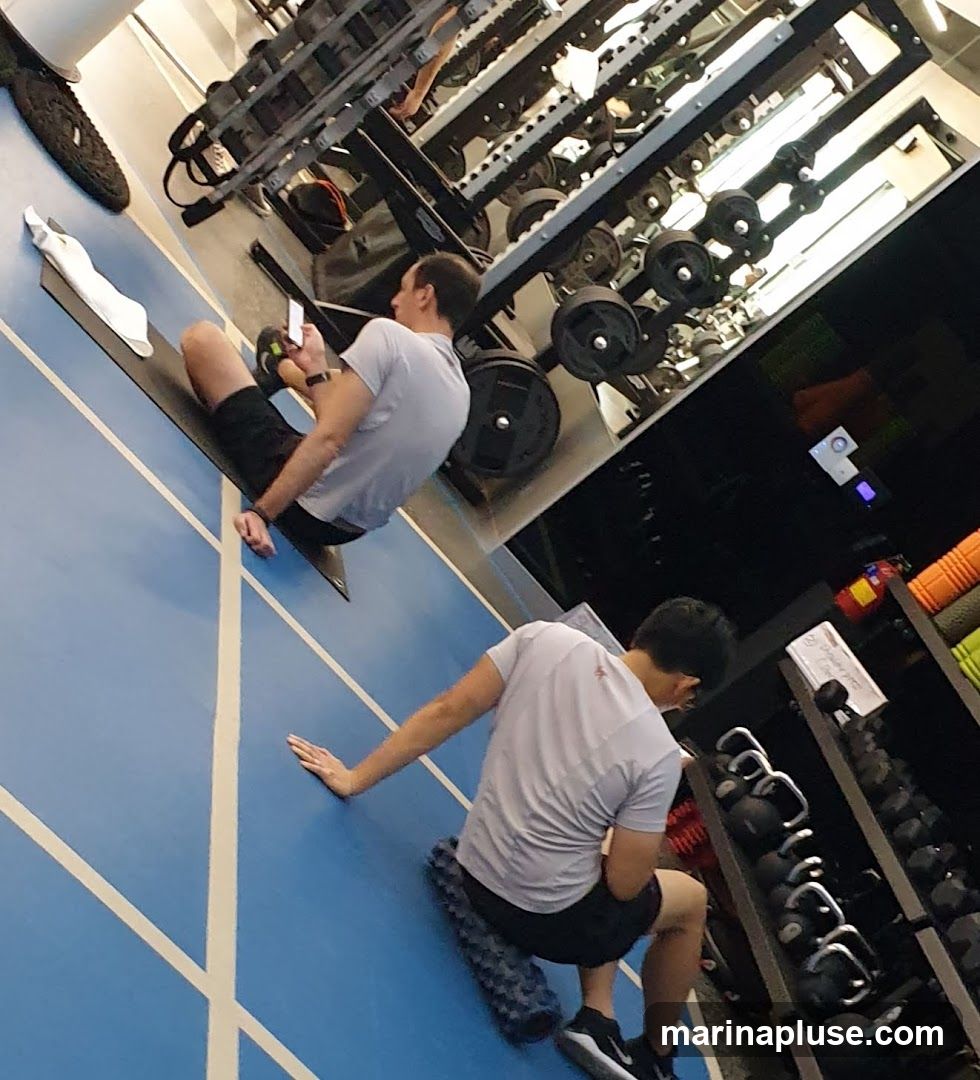 Two men are working out in a gym. One man is sitting on a mat and looking at his phone. The other man is sitting on a foam roller. There are weights and exercise equipment in the background.