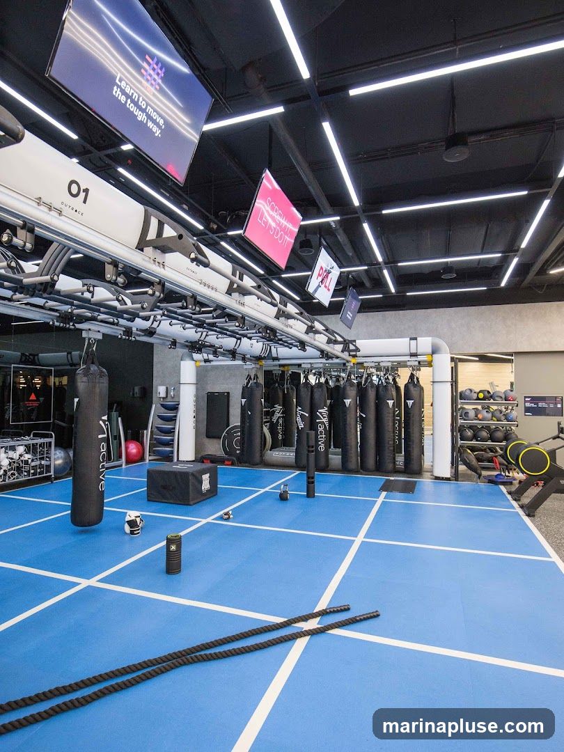 A spacious gym interior with a blue mat floor marked with white lines. Punching bags hang from the ceiling, and various workout equipment, including weights, ropes, and a plyometric box, are visible. Digital screens display motivational messages.