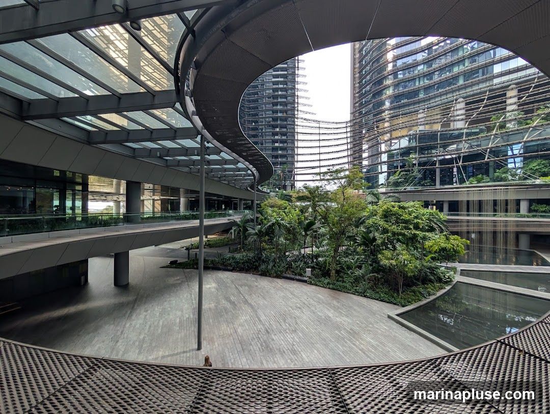 A wide-angle view of a contemporary outdoor space featuring a curved walkway with a glass roof, surrounded by modern buildings with reflective glass facades. Lush tropical plants and trees are integrated into the design, with several water features adding to the serene atmosphere. The ground is paved with wooden planks, and a lone figure is visible in the distance.