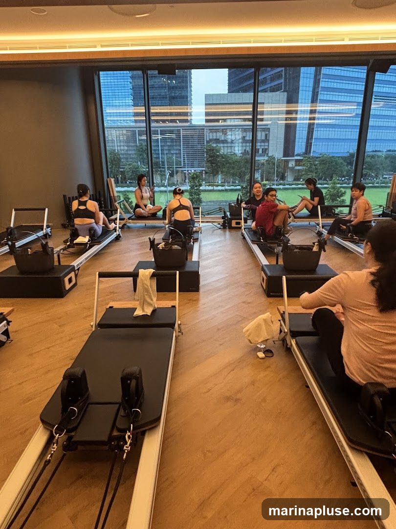 A group of people are sitting on Pilates reformers in a studio with large windows overlooking a city skyline. The room has wooden floors and modern lighting.