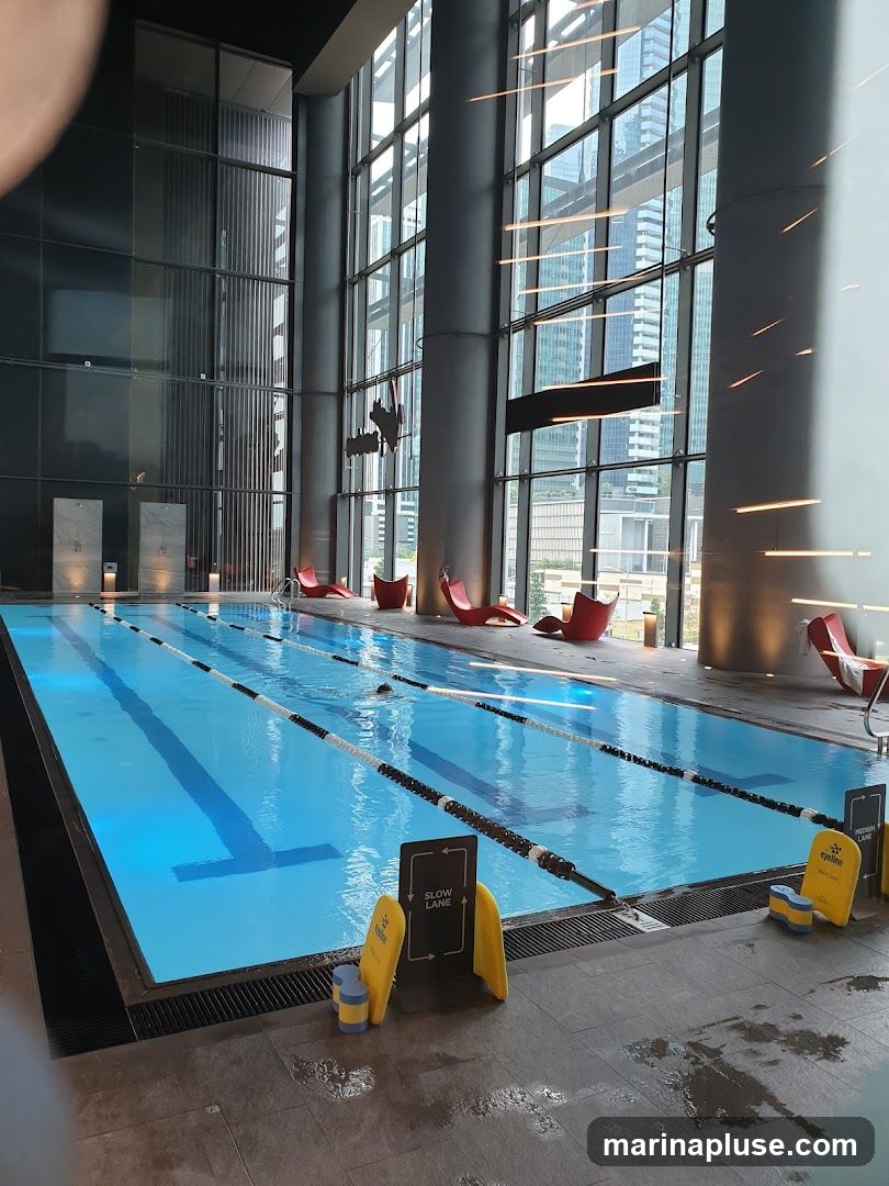 An indoor swimming pool with multiple lanes is illuminated by blue lights. The pool area features large windows with a view of city skyscrapers. Red lounge chairs are placed around the pool. A sign indicating 'SLOW LANE' is visible near the edge of the pool.