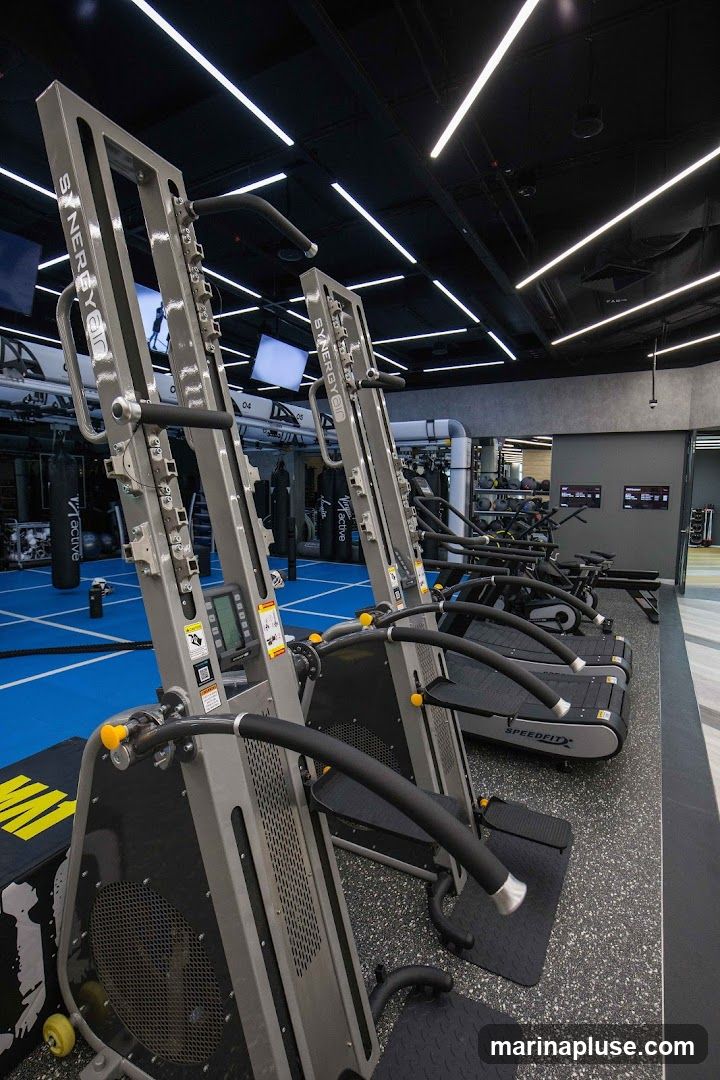 A row of vertical climbing machines and treadmills in a modern gym. The machines have the brand name "SYNERGYAIR" visible. The floor is a mix of blue matting and speckled grey rubber. In the background, punching bags and other gym equipment are visible.