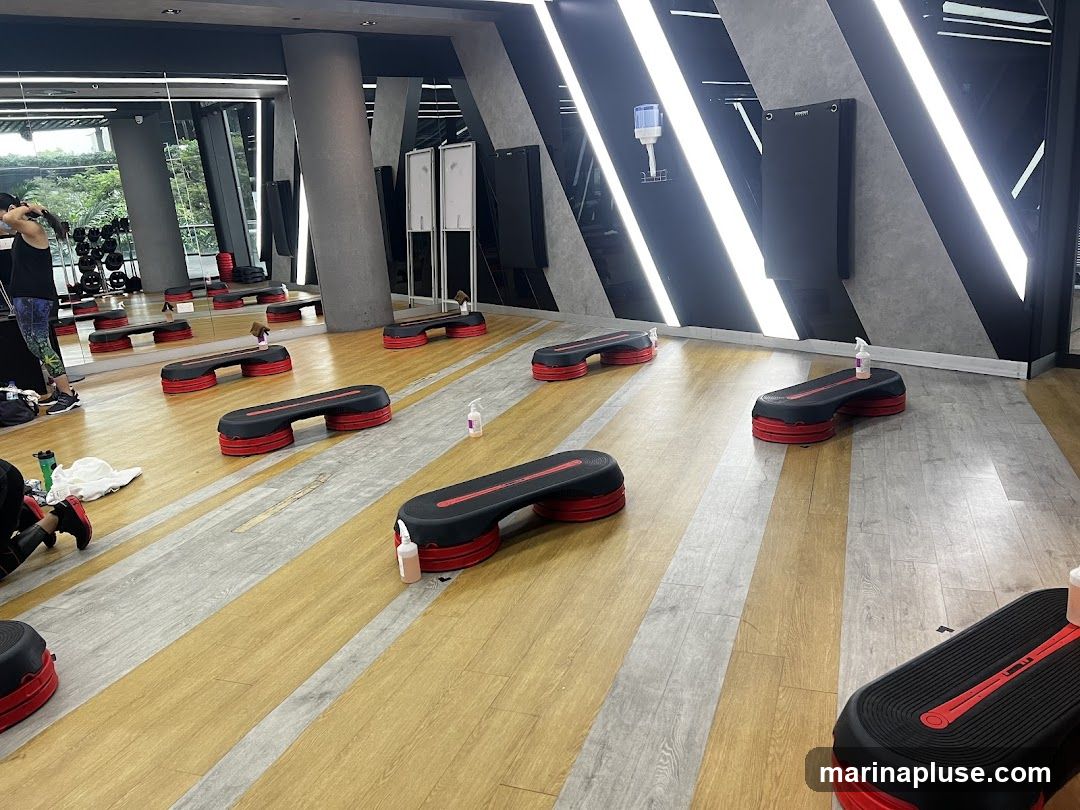 A gym studio with aerobic steps arranged for a class. The room has a wooden floor with grey and yellow stripes. Mirrors line one wall, reflecting exercise equipment and a person preparing for a workout. The other wall features a modern design with black panels and diagonal white LED lighting. Hand sanitizer bottles are placed on some of the steps.