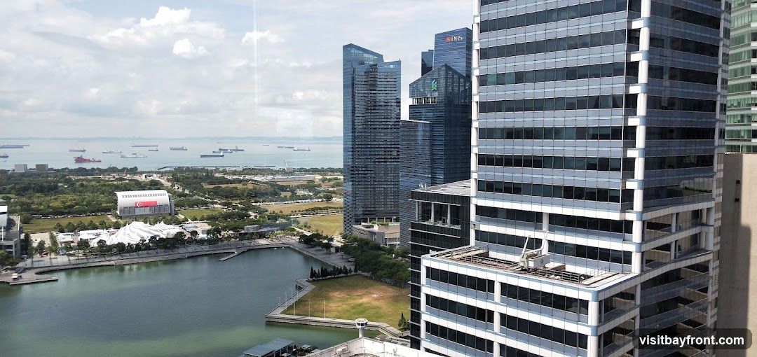 An aerial view of a modern cityscape featuring numerous skyscrapers and a busy harbor filled with ships. In the foreground, a large body of water is visible, with a park-like area and a distinctive white-domed structure on its edge. The background shows a vast expanse of the sea dotted with cargo ships under a cloudy sky. The architecture of the buildings suggests a financial or business district.