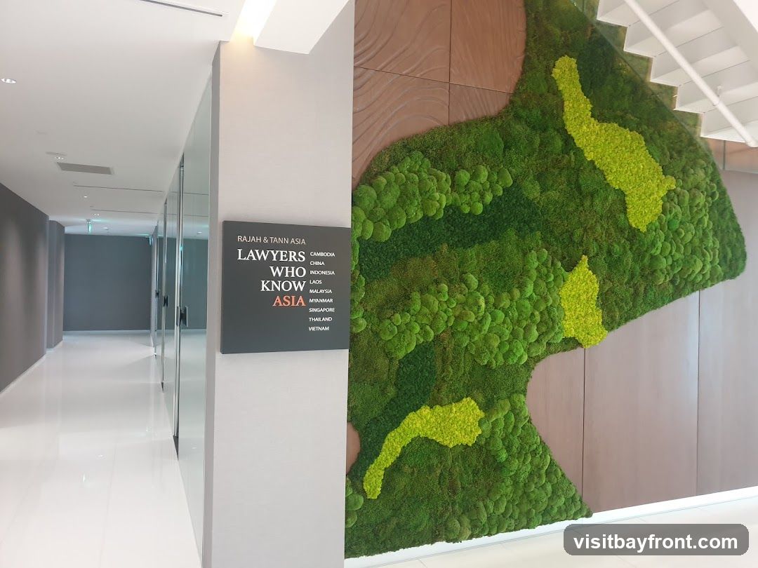 An interior view of a modern office hallway. On the right, a large, abstract art piece made of green moss is mounted on a wall. To the left of the moss art, a dark gray sign displays "RAJAH & TANN ASIA LAWYERS WHO KNOW ASIA" along with a list of Asian countries. The hallway is brightly lit with a white floor and gray walls, with glass-enclosed offices visible on the left.