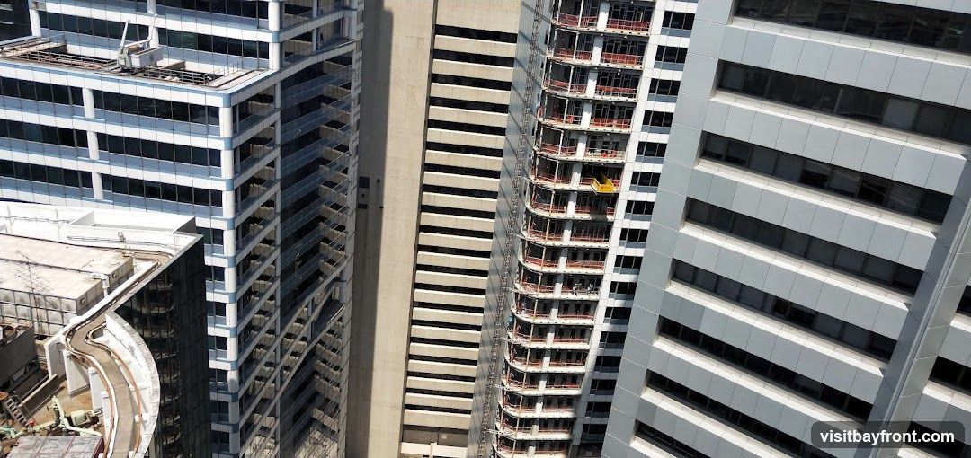 An aerial view shows several modern skyscrapers in a city. One building on the left has a white facade with blue trim and many windows. To its right, a building under construction shows its concrete structure and balconies. Further to the right, a building with a light gray facade and horizontal window bands is visible. Another building on the far right also features a gray facade with horizontal elements. The image captures the dense urban landscape from a high vantage point.