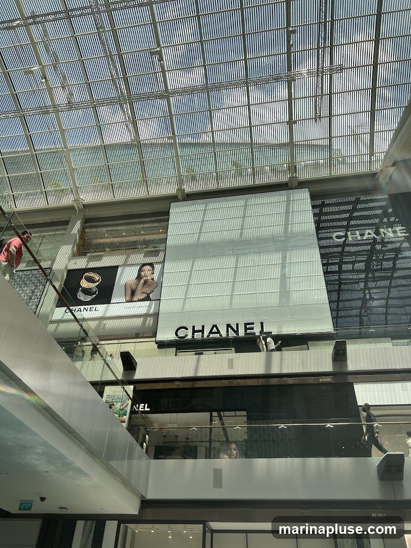 A wide shot of a multi-level shopping mall interior with a prominent Chanel store. The Chanel logo is visible on a large white facade above a darker storefront. There are also advertisements featuring jewelry and a model with the text "CHANEL COCO CRUSH" on the upper level. People are seen walking on the escalators and walkways. The ceiling is a modern glass and metal structure allowing natural light to filter in.