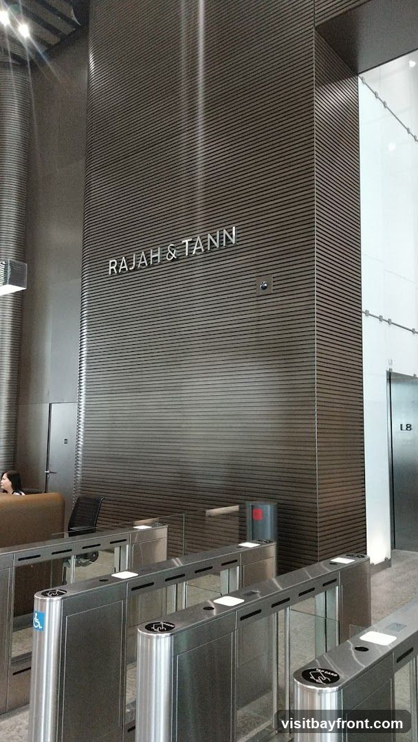 The image shows the interior of a modern office building lobby. A sign with the text "RAJAH & TANN" is visible on a wood-paneled wall. In the foreground, there are several metal turnstiles, some with accessibility symbols. An elevator bank is visible to the right, with a sign indicating "L8". A reception desk with a person is partially visible on the left.