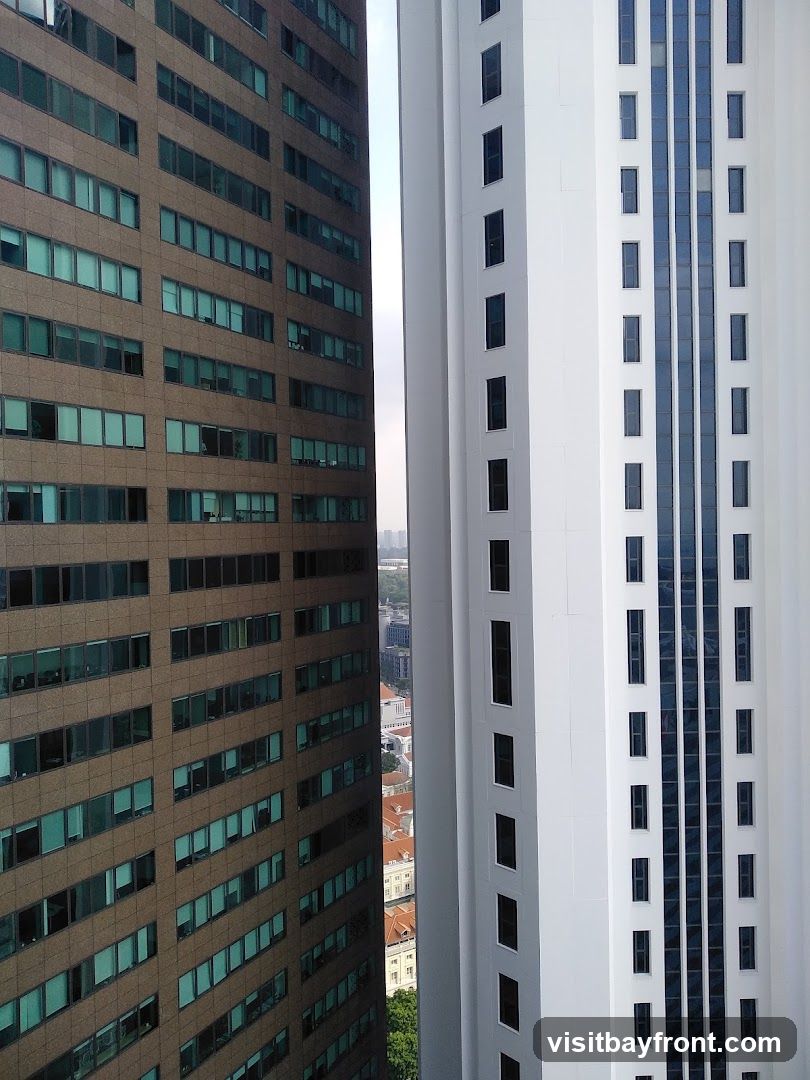 A view between two modern skyscrapers, one with a brown facade and numerous windows, and the other with a white facade and vertical blue-tinted windows. In the distance, a cityscape with lower buildings and a hint of greenery is visible.