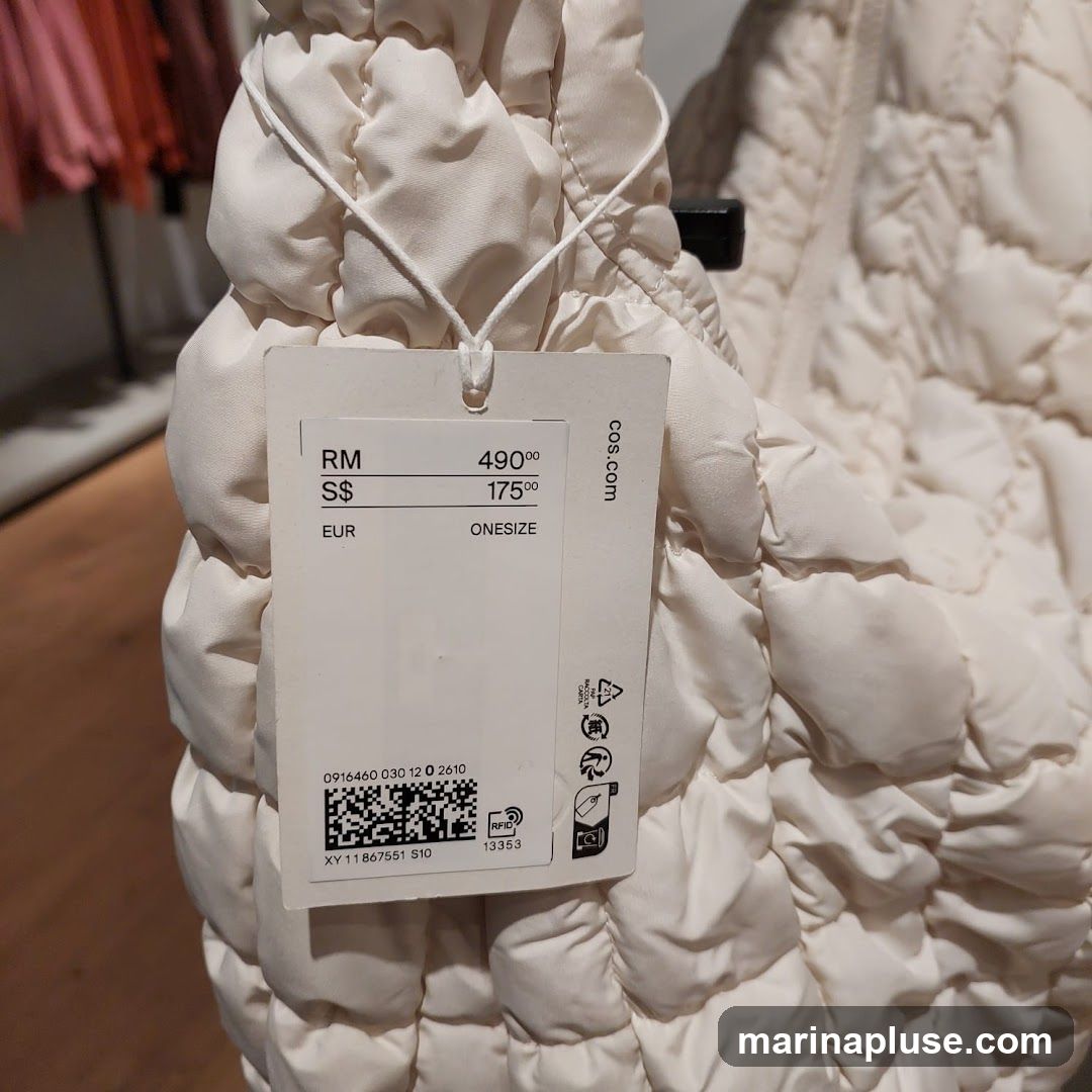 A close-up of a white product tag attached to a cream-colored quilted garment. The tag displays pricing information in multiple currencies: RM 490.00, S$ 175.00, and EUR. It also indicates the size as ONESIZE. The tag includes a barcode, RFID information, and the website "cos.com". In the blurred background, racks of clothing in pink and red hues are visible.