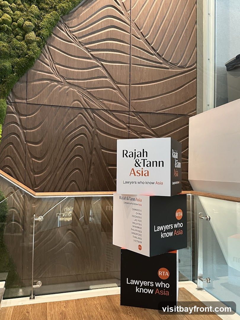Three stacked cubes with the Rajah & Tann Asia logo and slogan. The top cube is white with black text. The middle cube is black with white text and a list of Asian countries. The bottom cube is black with white text. The background features a textured wooden wall with a moss accent and a glass railing.