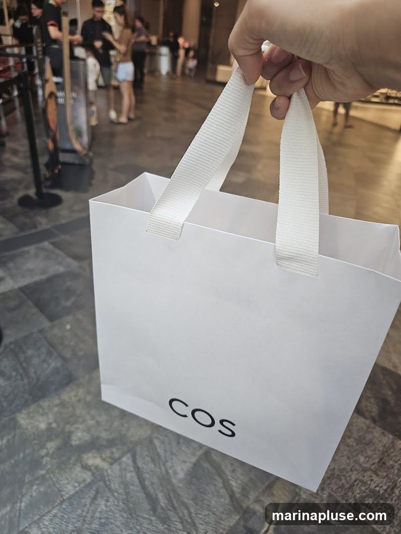 A white shopping bag with the COS logo printed in black is held by a hand. In the background, people are seen walking in what appears to be a shopping mall or retail environment.
