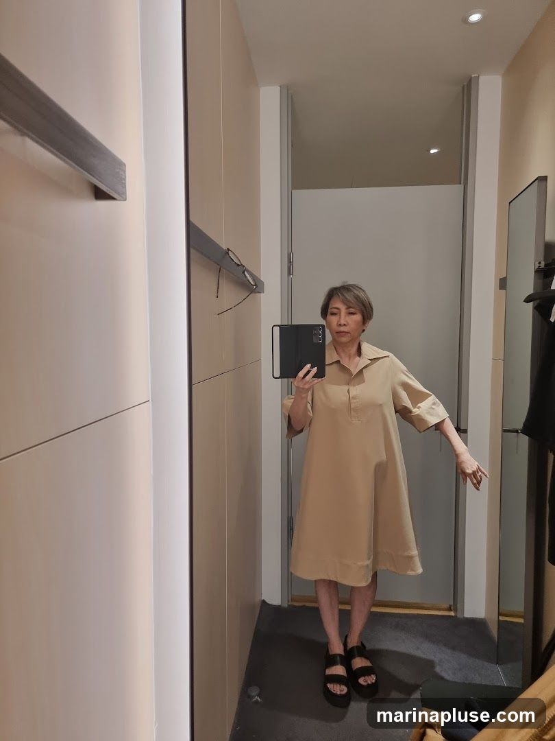 A woman stands in a fitting room, taking a mirror selfie. She is wearing a loose-fitting, knee-length beige dress with short sleeves and a collared neckline. She has on black platform sandals. The fitting room has light-colored walls, a mirror, and a clothing rack visible on the right.