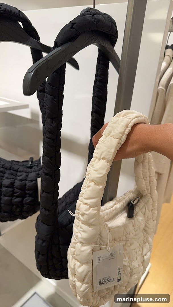 A hand holds a cream-colored puffy handbag with a ruched strap. Behind it, a black puffy handbag hangs from a wooden hanger. The handbags are displayed in a retail setting with other clothing items visible in the background. A price tag is attached to the cream handbag.