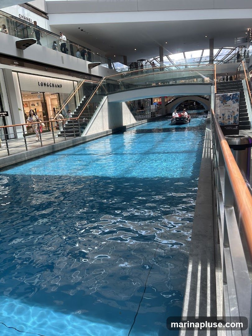 An indoor canal with bright blue water flows through a modern shopping mall. A gondola with two people navigates the waterway under a bridge. Shops like Chanel and Longchamp are visible on the upper levels, with escalators and staircases connecting the floors. Sunlight streams in from the ceiling, illuminating the scene.