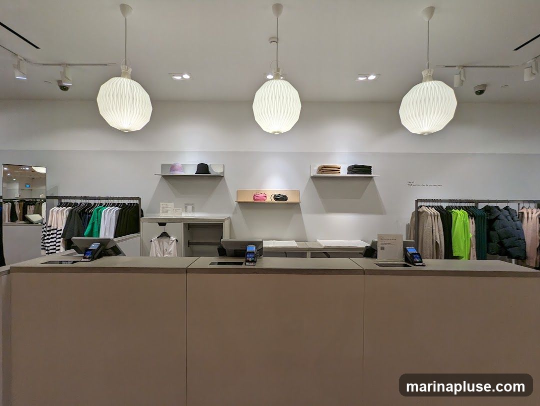 A view of a clothing store's interior, showing a checkout counter in the foreground and racks of clothing in the background. Three large, pleated pendant lights hang from the ceiling. On shelves above the counter are hats and folded clothing. Racks display various garments, including sweaters, jackets, and shirts in a range of colors. A sign on the wall reads "Like it? What just it in a bag for you here.". There are also security cameras visible on the ceiling.