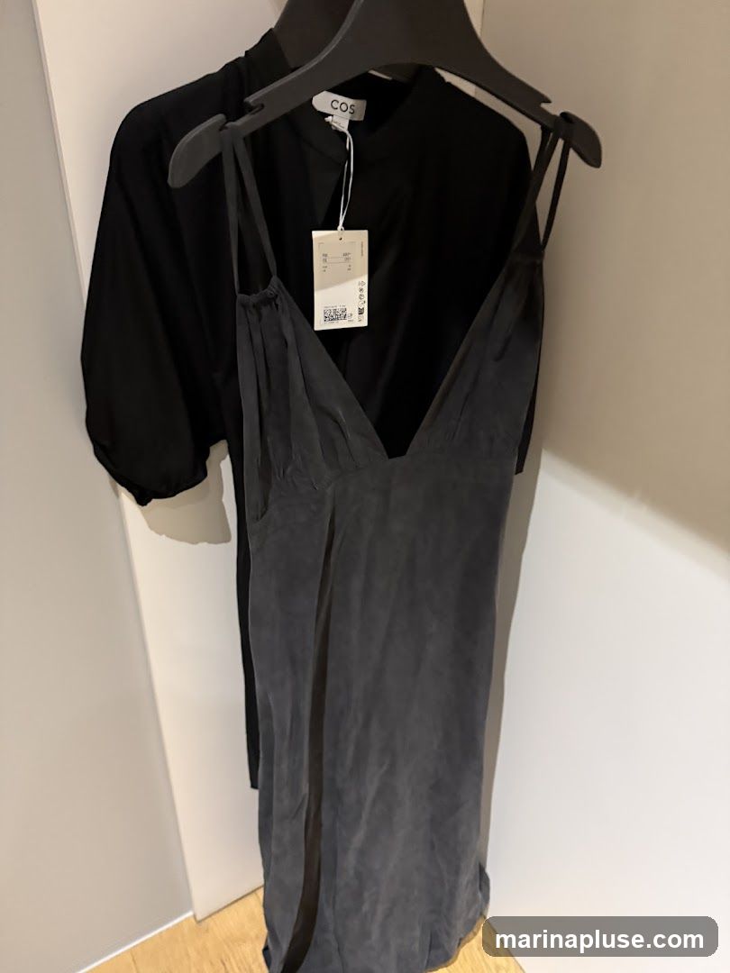 A dark grey slip dress with thin straps hangs on a hanger in front of a black garment. A white tag with the COS logo is visible on the black garment.