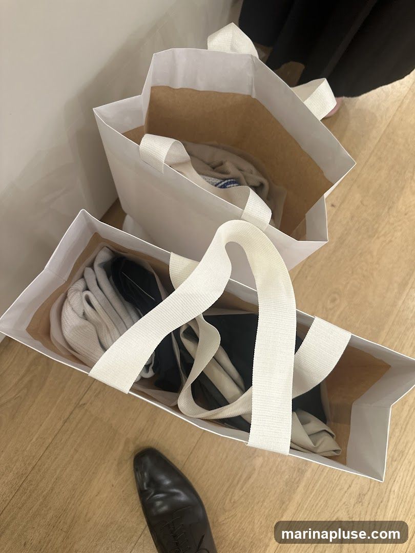 Two white paper shopping bags with white handles are filled with folded clothing. A black dress shoe is visible in the foreground on a wooden floor.