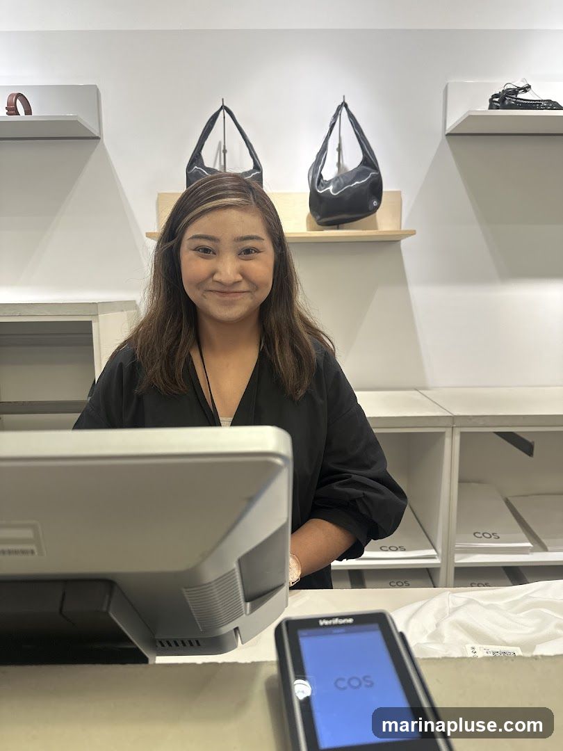 A smiling sales associate stands behind a checkout counter in a retail store. The store appears to sell handbags and shoes, with several bags displayed on shelves behind her. A payment terminal with the COS logo is visible on the counter.