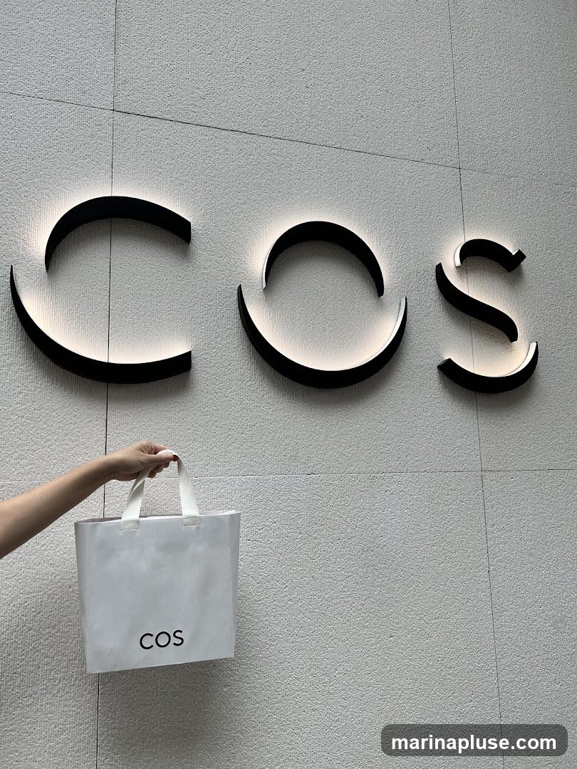 A hand holds a white shopping bag with the COS logo. Behind it, the COS logo is displayed as illuminated black lettering on a textured wall.