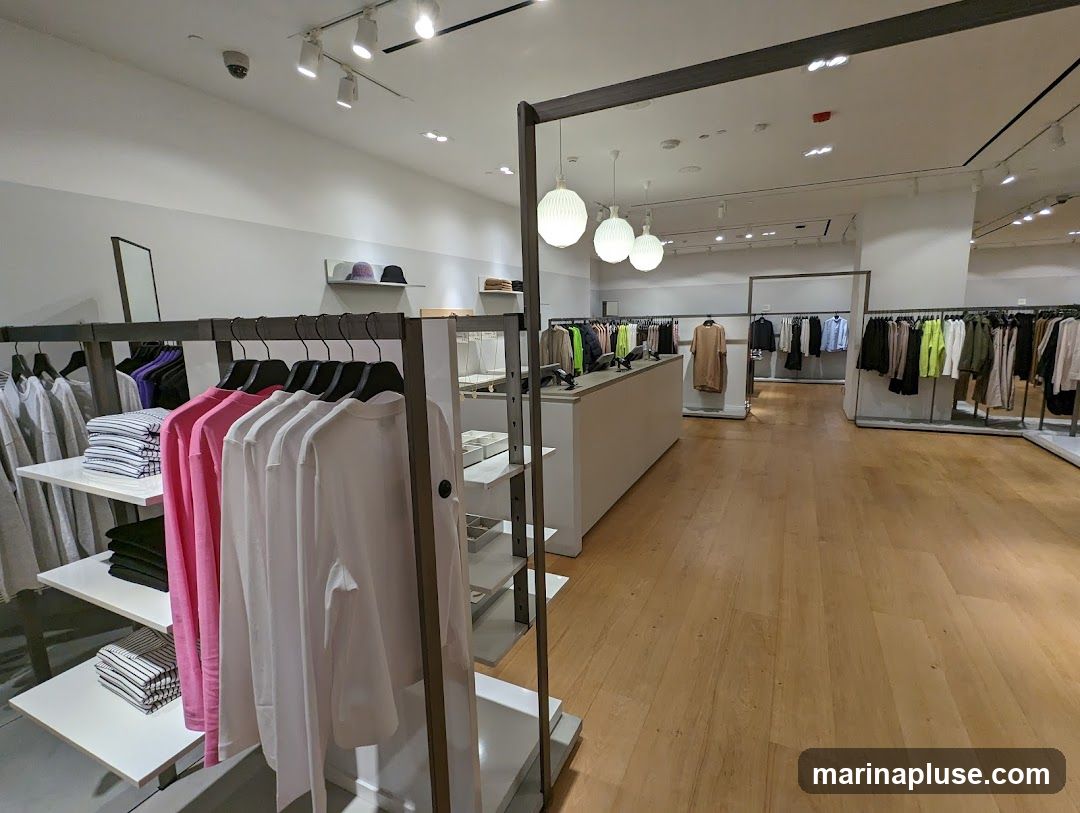 A well-lit clothing store interior with racks of apparel. In the foreground, there are shelves with folded striped and solid-colored shirts, and hanging long-sleeved shirts in pink and white. Further into the store, more clothing racks are visible, displaying a variety of garments. A long counter with what appears to be a cash register or display screens is situated in the middle ground. The flooring is light-colored wood.