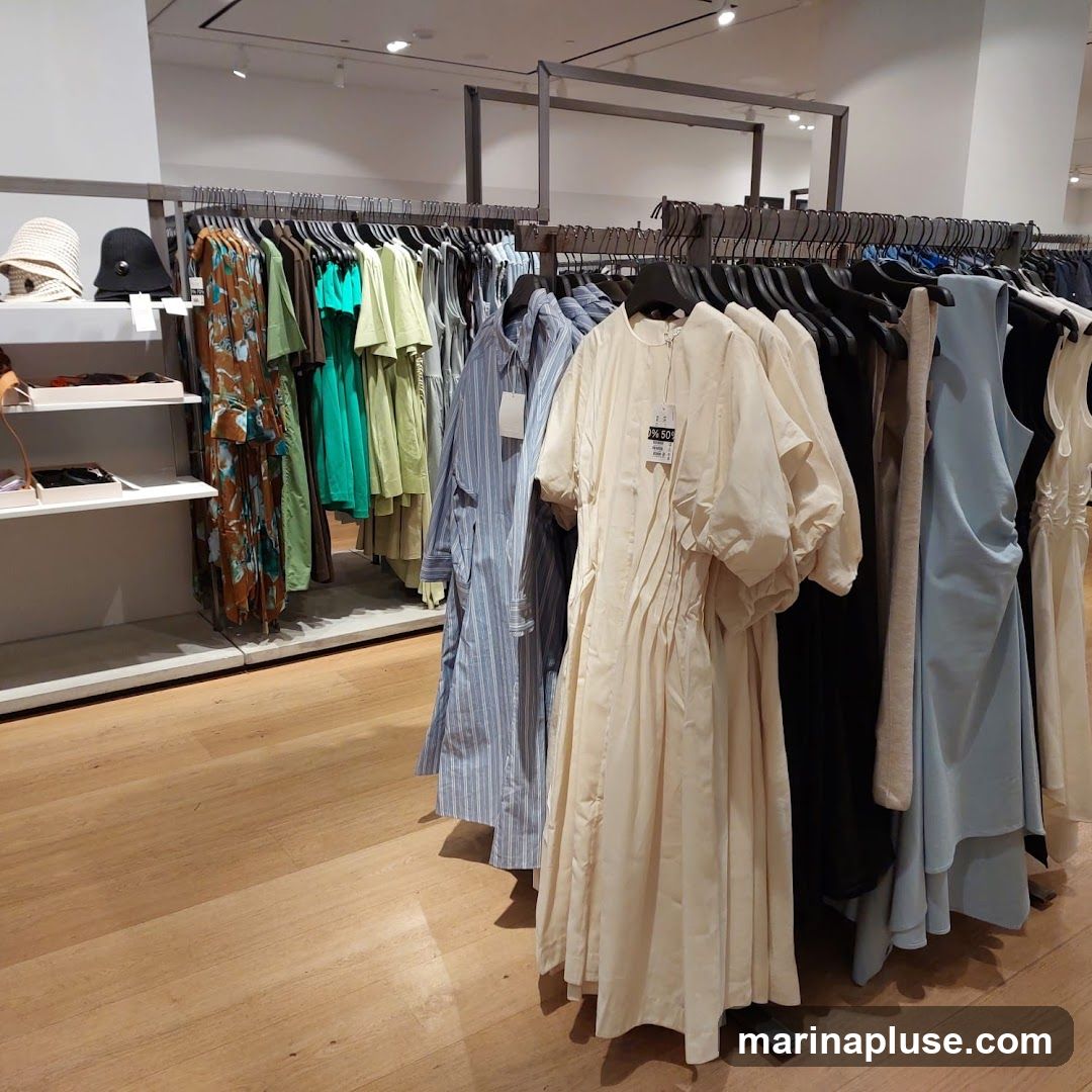 A clothing store interior with racks of dresses and other garments. On the left, shelves display hats and accessories. The garments on display include patterned dresses, green and lime green tops, blue striped dresses, and a cream-colored dress with puffed sleeves. A tag on the cream dress indicates a 50% discount. The store has a wooden floor and bright overhead lighting.