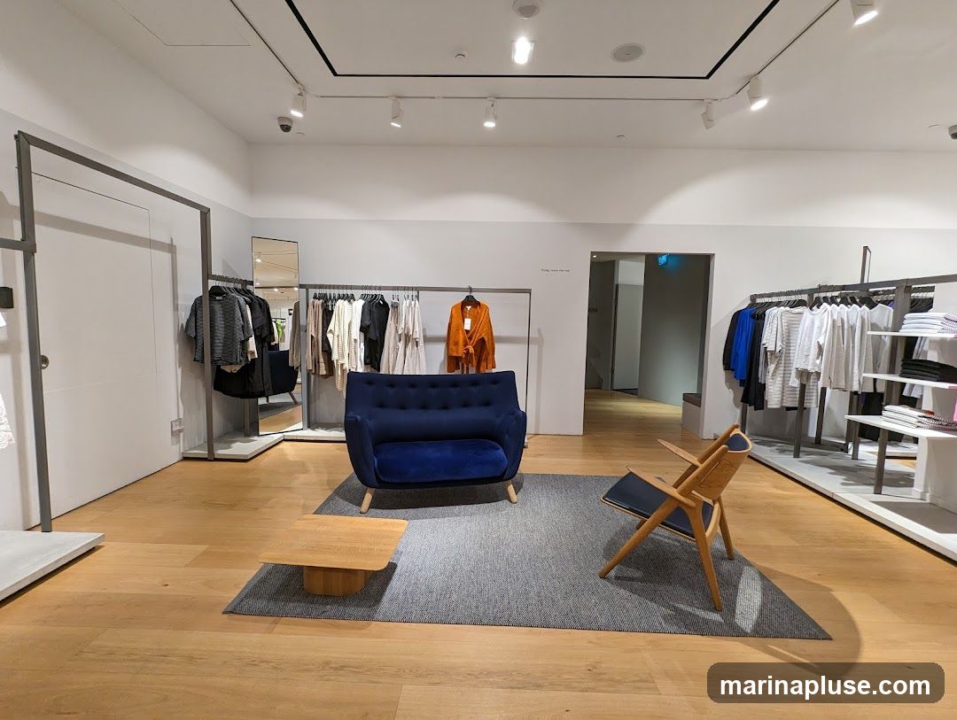 A well-lit clothing store interior featuring racks of apparel, a seating area with a blue sofa and a wooden chair, and a coffee table. The floor is made of light-colored wood, and the walls are white. A doorway leads to another part of the store.